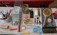 Overhead view showing entire assortment: cage trap, bug zapper racket, shears, scissors, saw blades, pest traps, tape, trimmer line, cable ties, multi-tool, brush, sharpening block, telescopic rod, screws box