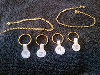 Four rings with size tags and two gold-tone chains displayed on dark fabric