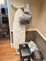 Ironing board with attached white cover, loose grey cover draped, liners and trash bins visible