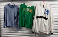Front view of three shirts hanging: blue long sleeve, green Ireland T-shirt, white hooded windbreaker
