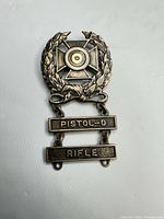 Front view of sterling silver marksmanship medal showing wreath, target cross, and PISTOL-D & RIFLE dangle bars