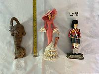 Three figurines aligned: cat with mice, lady with parasol, Scottish soldier; tape measure shows height reference