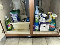 Overall view under sink showing Ajax dish soap, dishwasher pods canister, Febreze aerosol, several spray cleaners, PUR filter box