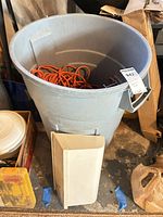 Trash can holding orange extension cord, wall-mount box leaning against it