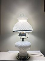 Full view of lamp lit showing milk glass hobnail base, shade, brass accents and chimney