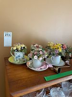 Group view of four teacup floral arrangements and two brass watering cans
