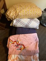 Stack of bedding showing yellow floral/stripe covered duvet, white check comforter, purple comforter, pink dragon duvet cover