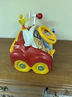 Side view of red and yellow dashboard toy showing steering wheel, Big Bird figure, horn, lever and twin yellow wheels with blue B logo