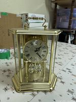 Front view of Bulova brass-tone anniversary mantel clock showing dial, rotating pendulum, glass panels, top handle, and commemorative tag on base