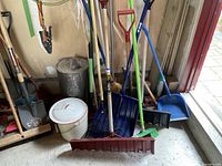Group of mixed long-handled tools, buckets and cleaning items leaning against garage wall