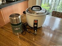 Rice cooker with lid and power cord beside Stanley thermos