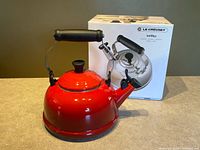 Front view of red Le Creuset tea kettle next to original box