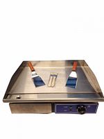 Front view of stainless steel electric griddle with two spatulas and two brushes on cooking surface, showing control panel