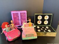 Group shot showing three dolls seated in two pink cars, wardrobe, and closed tea set box
