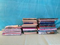 Three stacks of assorted cookbooks, spiral-bound recipe books, and magazines
