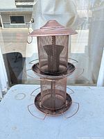 Full view of copper-tone metal mesh bird feeder showing lid, mesh body, two trays and hanging loop