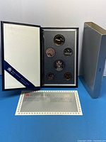 Open blue presentation book displaying six coins, COA card, and damaged grey sleeve visible