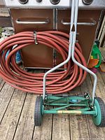 Coiled red garden hose and green Lee Valley reel mower in front of barbecue
