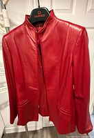 Front view of red collarless leather jacket on hanger showing slit pockets and zip front