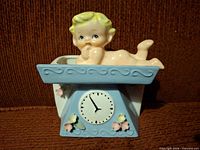 Front view showing baby figure atop light blue scale face with flowers and clock dial