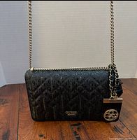 Front view of Guess black coated canvas crossbody bag showing monogram pattern, flap, gold logo plaque and chain strap