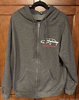 Front view of dark gray zip hoodie on hanger showing graphic, pockets, zipper, drawcord