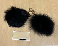 Two black fur pom-pom keychains with tags on wooden surface