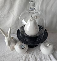 Group shot showing all items: cloche on black stand with pear inside, rabbit figurine, two apples