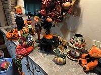 Countertop view showing multiple pumpkin-head figurines, seated scarecrow doll, ceramic lamp, plush bear, frame and resin pumpkin