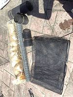 Multiple black plastic mesh rolls, galvanized wire roll, burlap roll in plastic, and flat mesh sheet on paved surface