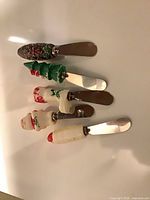 Overhead view of five holiday-motif cheese spreaders showing stainless blades and resin handles