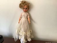 Full view of doll standing against wall wearing ivory lace gown