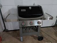 Front view of Weber Spirit II gas grill showing three burner knobs, side shelves, cart frame with one wheel