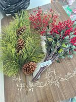 Bundle of faux pine needles with pine cones and bundle of red holly berry stems on tabletop