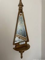 Full view of mirror showing triangular shape, gold frame, top and bottom finials, and shelf