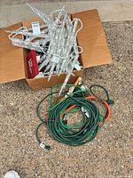 Box containing multiple clear LED icicle light strands next to coiled green and orange extension cords on ground