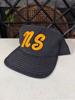 Front view of black snapback hat showing orange 'NS' embroidery and brim