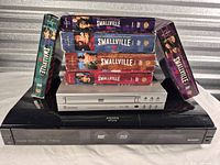 Sharp Aquos Blu-ray player, CyberHome DVD player stacked with six Smallville DVD season box sets