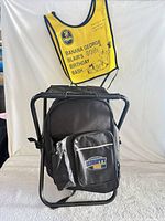 Front view of black backpack cooler on folding stool frame with yellow Banana George bib hanging above