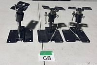 Three identical black adjustable speaker brackets standing upright on table, showing base plates, swivel joints, safety wires