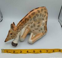 Left side view of porcelain giraffe showing head down, legs folded, ruler indicates length