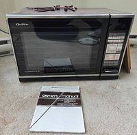 Closed microwave displaying exterior, power cord on top, owner’s manual in front