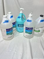 Front view of five gallon-size bottles: four white hand sanitizers and one blue Hibiclens skin cleanser