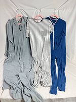 Three onesies/jumpsuits hanging: gray fleece hooded, gray-white striped, blue jersey