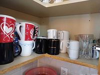 Assortment of mugs on shelf including red heart mugs, black Road Garage mugs, white blue-band mugs and others