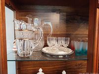 Clear pressed glass pitcher beside stacked glasses and silver-tone tray with cordials on glass shelf