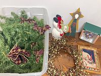 Overview of all items: evergreen garland with bows in plastic bin, snowman figurine, wooden angel, pip-berry garland, wooden snowman plaque and boxed ornament