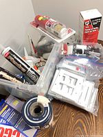 Mixed handyman supplies in clear bin showing adhesive tubes, drywall tape roll, cover plates