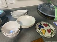 Stack of three white mixing bowls, five white oblong bowls, five white shallow spaghetti bowls and one fruit motif plate displayed on countertop
