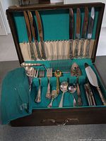 Open wooden cutlery chest showing assorted silverplate flatware and serving pieces in teal interior